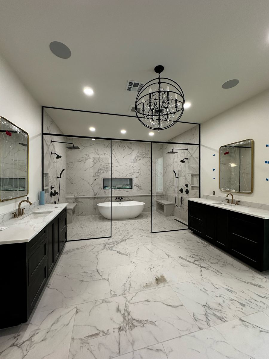 Project Pilot project: Large primary bathroom with a black-framed glass wet room containing a freestanding tub and two shower heads, dark dual vanities, marble-look floor tile, and a chandelier