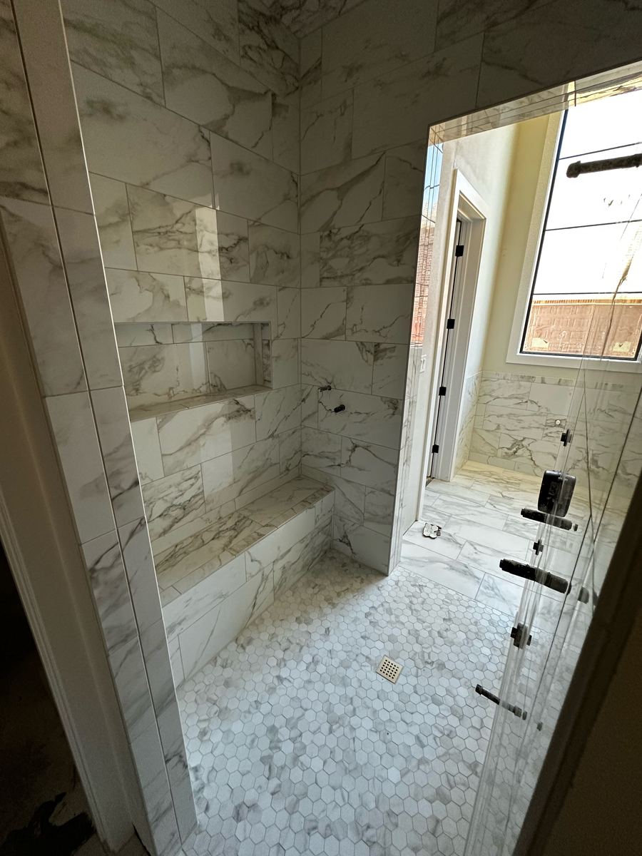 Project Pilot project: Marble-look tiled walk-in shower with a built-in bench, recessed niche, white hexagon floor tile, and a frameless glass door